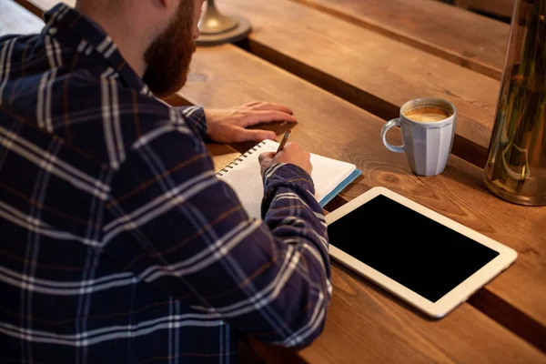 Joven hombre de negocios barbudo se sienta en la cafetería, casa en la mesa y escribe en el portátil, cerca se encuentra el ordenador tableta con pantalla negra. El hombre está trabajando, estudiando . — Foto de Stock