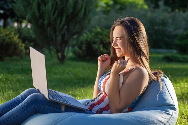 Молодая симпатичная девушка с ноутбуком, сидит на фасолевом пакете в саду или парке, на зеленой траве. Концепция покупок — стоковое фото