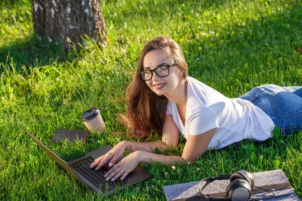 Молодая женщина использует ноутбук в парке, лежащий на зеленой траве. Концепция досуга. — стоковое фото