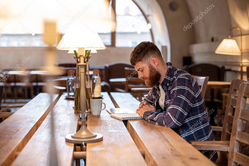 Young bearded businessman sits in cafe, home at table and writes in notebook, near lies tablet computer. Man is working, studying.