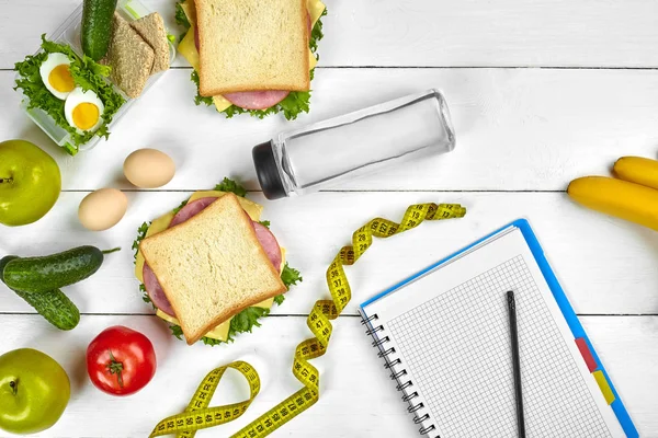 Üstten görünüm defter ile kopya alanı ve sağlıklı kahvaltı planlama. Jambon ve peynir, yumurta, salatalık, domates, Beyaz ahşap masa su şişe ile sandviç.