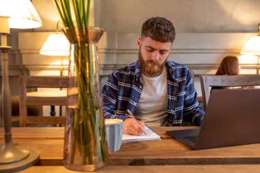 Geçici iş adamı veya serbest meslek sahibi eserleri üstünde defter, planlama ile akıllı telefon, kahve kahve dükkanı veya ev ofis, masada dizüstü bilgisayar üzerinde çalışan.