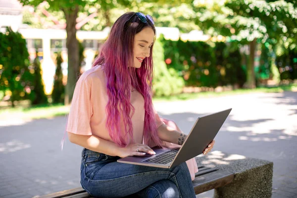 Молодая девушка учится в весеннем парке, сидит на деревянной скамейке и просматривает ноутбук — стоковое фото