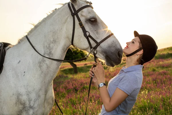 Жокей молодая девушка ласкает и обнимает белого коня на закате — стоковое фото
