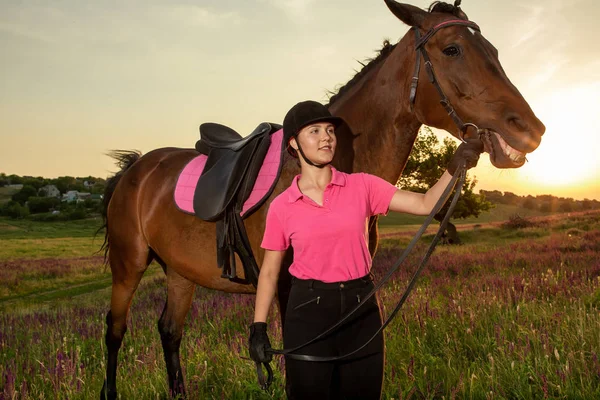 Beautiful smiling girl jockey stand next to her brown horse wearing special uniform on a sky and green field background on a sunset.