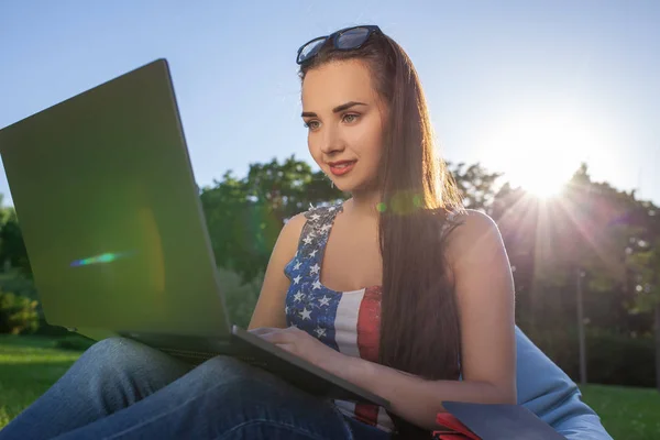 Красивая молодая женщина, сидящая на бобах мешок использовать ноутбук во время отдыха на траве в парке на солнце — стоковое фото
