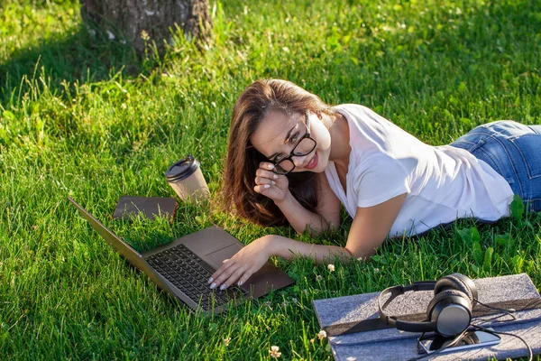 Jovem mulher usando laptop no parque deitado na grama verde. Conceito de atividade de lazer. — Fotografia de Stock