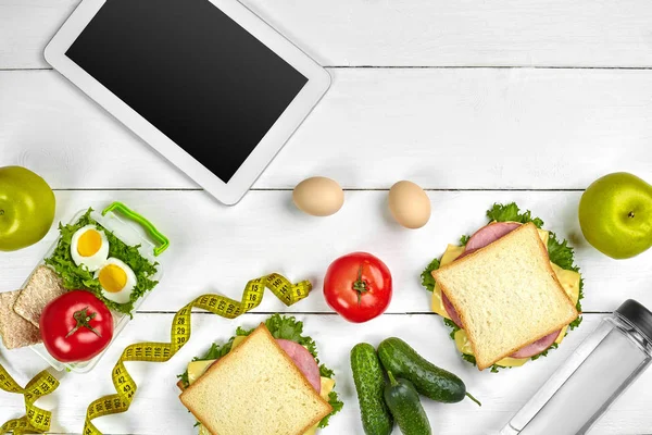 Witte tablet pc met een leeg scherm op de keukentafel met broodjes, verse groenten, fles water en apple. Lunch — Stockfoto