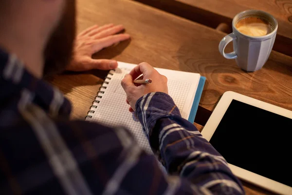 Joven hombre de negocios barbudo se sienta en la cafetería, casa en la mesa y escribe en el portátil, cerca se encuentra el ordenador tableta con pantalla negra. El hombre está trabajando, estudiando . — Foto de Stock