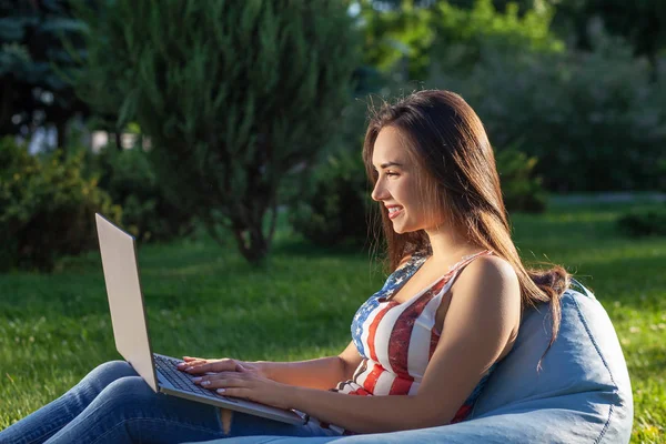 Jeune fille mignonne avec ordinateur portable, assis sur un sac de haricots dans le jardin ou le parc, sur l'herbe verte. Concept d'achat en ligne — Photo