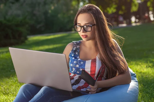 Молодая симпатичная девушка с ноутбуком, сидит на фасолевом пакете в саду или парке, на зеленой траве. Современный образ жизни — стоковое фото