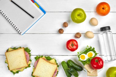 Üstten görünüm defter ile kopya alanı ve sağlıklı kahvaltı planlama. Jambon ve peynir, yumurta, salatalık, domates, Beyaz ahşap masa su şişe ile sandviç.