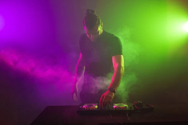 DJ en el trabajo mezclando sonido en sus cubiertas en una fiesta o club nocturno con un colorido fondo de luz de humo — Foto de Stock