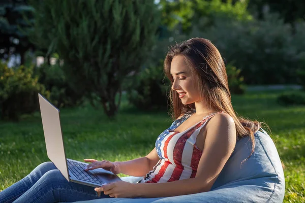 Jeune fille mignonne avec ordinateur portable, assis sur un sac de haricots dans le jardin ou le parc, sur l'herbe verte. Concept d'achat en ligne — Photo