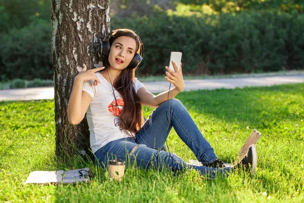 Woman relax with headphones listening to music sitting on grass in park. Young woman with headphones doing selfie on her smartphone