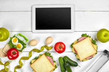 Mutfak masasında sandviç, taze sebze, elma ve su şişe ile boş bir ekranda beyaz tablet bilgisayar. Öğle Yemeği