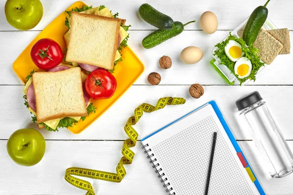 Bovenaanzicht planning notitieboekje met kopie ruimte en gezond ontbijt. Sandwich met ham en kaas, eieren, komkommer, tomaat en fles water op witte houten tafel. — Stockfoto