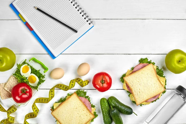 Top view planning notebook with copy space and healthy breakfast. Sandwich with ham and cheese, eggs, cucumber, tomato and bottle of water on white wooden table.