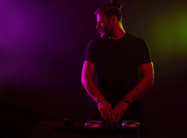 DJ en el trabajo mezclando sonido en sus cubiertas en una fiesta o club nocturno con un colorido fondo de luz de humo — Foto de Stock
