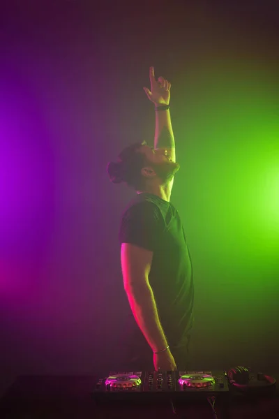 DJ en el trabajo mezclando sonido en sus cubiertas en una fiesta o club nocturno con un colorido fondo de luz de humo — Foto de Stock