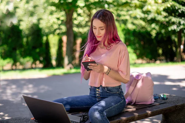 Женщина пишет в блокноте, сидя на деревянной скамейке в парке. Девушка работает на открытом воздухе на портативном компьютере, копировальное пространство . — стоковое фото