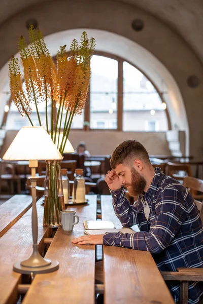 Young bearded businessman sits in cafe, home at table and writes in notebook, near lies tablet computer. Man is working, studying.