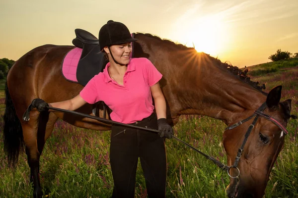Beautiful smiling girl jockey stand next to her brown horse wearing special uniform on a sky and green field background on a sunset. — Stock Photo, Image