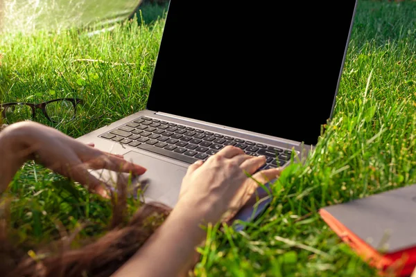 Close up hands on keyboard. Woman working on laptop pc computer with blank black empty screen to copy space in park on green grass sunshine lawn outdoors.