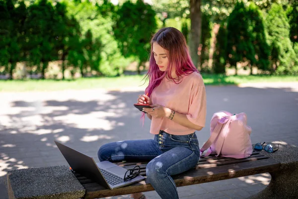 Женщина пишет в блокноте, сидя на деревянной скамейке в парке. Девушка работает на открытом воздухе на портативном компьютере, копировальное пространство . — стоковое фото