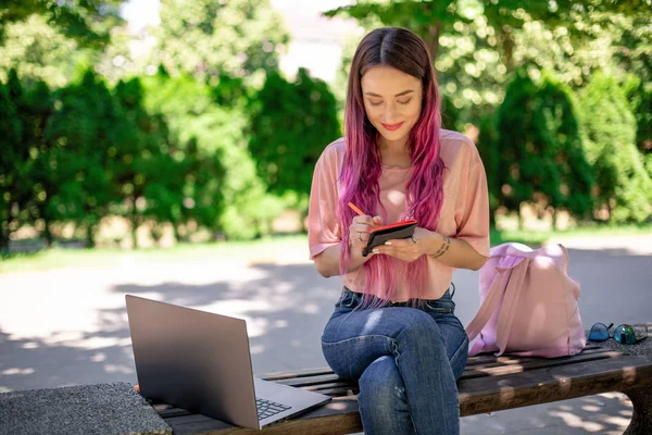 Femme écrivant dans un cahier assis sur un banc en bois dans le parc. Fille travaillant à l'extérieur sur ordinateur portable, espace de copie . — Photo