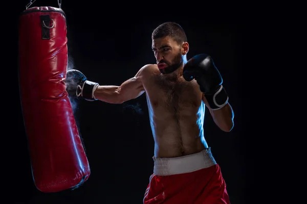 Entrenamiento de boxeador masculino barbudo con saco de boxeo sobre fondo negro. Boxeador masculino como ejercicio para la gran pelea . — Foto de Stock