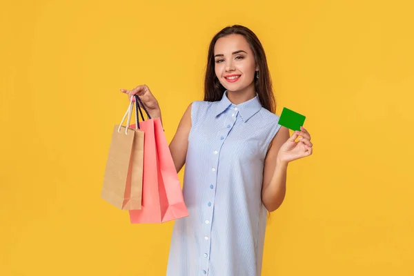 Mujer de moda de 20 años en vestido con el pelo largo marrón sonriendo mientras sostiene diferentes paquetes de compras y tarjeta en las manos aisladas sobre fondo amarillo — Foto de Stock