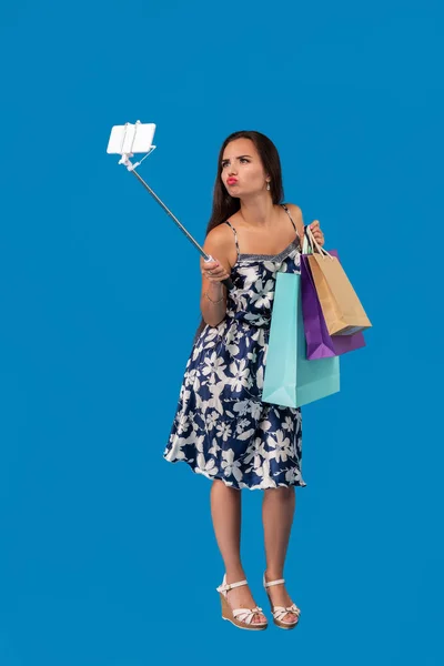 Poz ve alışveriş torbaları üzerinde mavi bir arka plan ile telefonda bir selfie alarak genç şık kadın — Stok fotoğraf