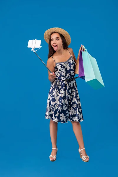Fiatal nő alkalmi ruhák a bevásárló szatyrok selfie stick segítségével vesz egy önarckép a kék stúdió háttér-val másol hely — Stock Fotó