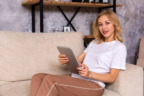 Szőke nő ül a kanapén használata tabletta otthon a nappaliban — Stock Fotó