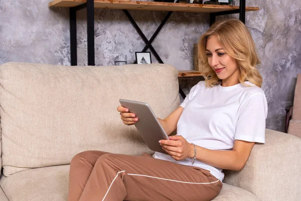 Szőke nő ül a kanapén használata tabletta otthon a nappaliban — Stock Fotó