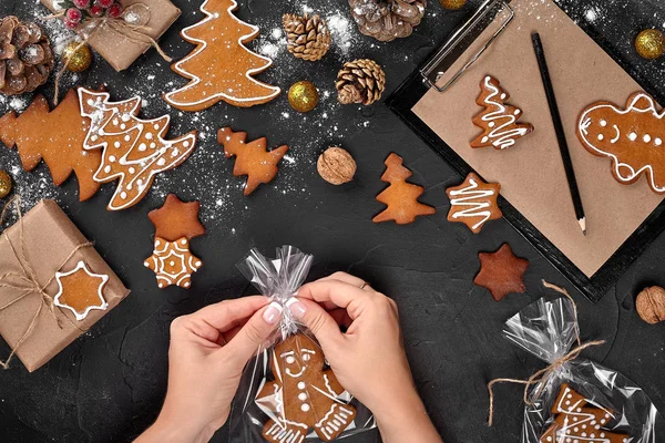 Christmas gift gingerbread on dark background. Biscuits in festive packaging. Woman is packaging Christmas gingerbread cookies with icing sugar. Top view — Stock Photo, Image