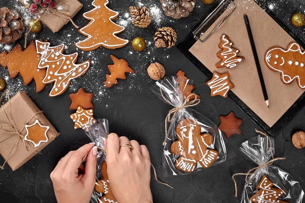 Pão de gengibre presente de Natal no fundo escuro. Biscoitos em embalagens festivas. Mulher está embalando biscoitos de gengibre de Natal com açúcar de confeiteiro. Vista superior — Fotografia de Stock