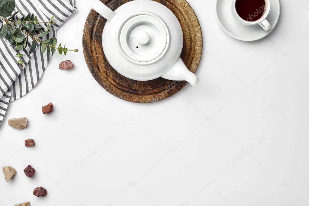 A white porcelain teapot on a wooden board and a white cup with tea on a table. Top view. Copy space.