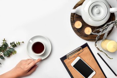 Genç kadın sağ üstünde çay bardağı, akıllı telefon ve su ısıtıcısı yanında beyaz masada boş defter sabah zamanında yazma