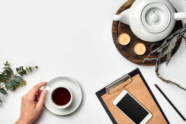 Joven mujer derecha escribiendo en un cuaderno en blanco en una mesa blanca con taza de té, teléfono inteligente y hervidor de agua al lado en la mañana — Foto de Stock
