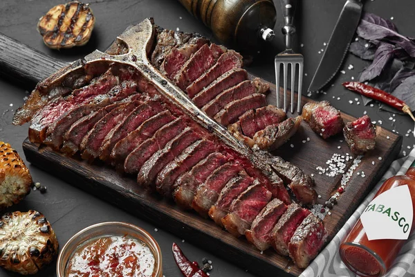 Bife em fatias T-bone deitado em tábua de madeira. Verduras grelhadas em uma grelha de panela. Mesa preta com pano cinza — Fotografia de Stock