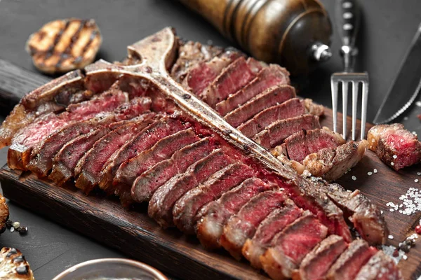 Bife em fatias T-bone deitado em tábua de madeira. Verduras grelhadas em uma grelha de panela. Fundo preto. Close-up — Fotografia de Stock