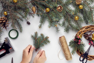 Kadın elinin üst görüntüsü Noel çelengi yapar. Paketlenmiş hediyeler ve parşömenler, ladin dalları ve aletler beyaz masada. Noel ya da Yeni Yıl tatiline hazırlanılıyor