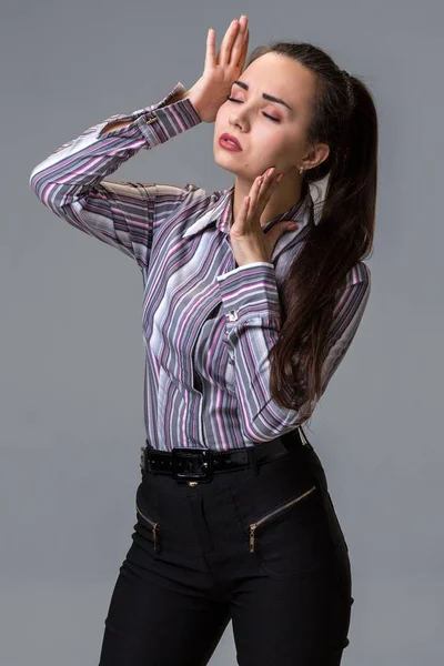 Studio portrait of brunette girl in striped shirt, trousers and long tail posing against grey background. — Stock Photo, Image