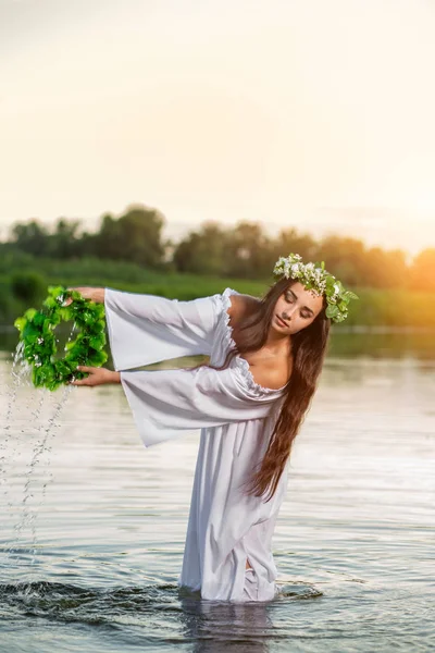 Beautiful black haired girl in white vintage dress and wreath of flowers standing in water of lake. Sun flare.