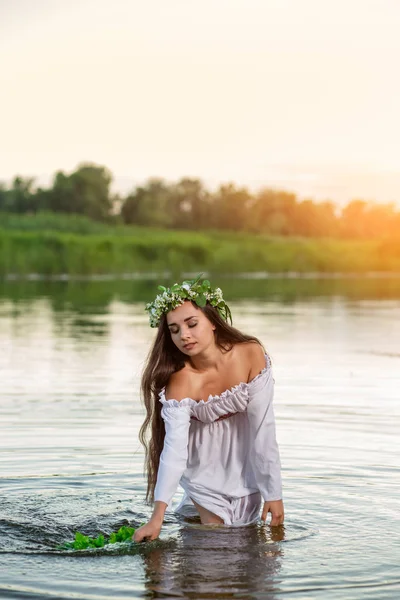Güzel siyah saçlı kız beyaz vintage elbise ve göl suda ayakta çiçek çelenk. Güneş parlaması.