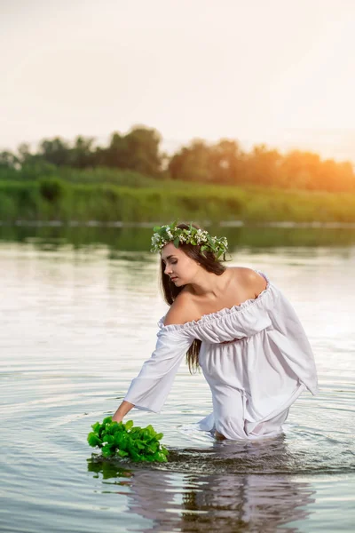 Beautiful black haired girl in white vintage dress and wreath of flowers standing in water of lake. Sun flare.