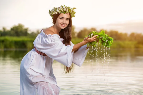 Beautiful black haired girl in white vintage dress and wreath of flowers standing in water of lake. Sun flare.