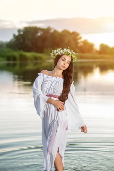 Beautiful black haired girl in white vintage dress and wreath of flowers standing in water of lake. Sun flare.